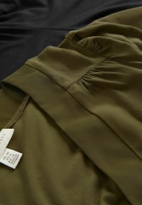 Olivgrüner Stoff mit weicher Textur und Raffung, neben einem glatten schwarzen Stoff platziert. Pflegeetiketten sind auf dem olivfarbenen Kleidungsstück sichtbar.