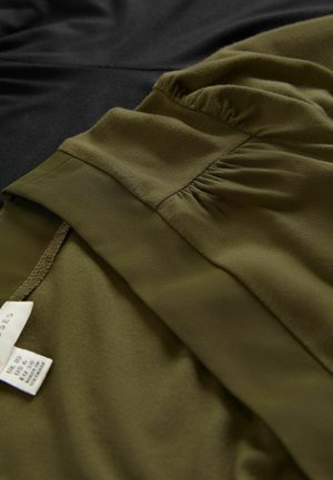 Oliivinvihreä kangas, jossa on pehmeä rakenne ja poimuttunut yksityiskohta, sijoitettu sujuvasti mustan kankaan viereen. Huolenpitotarrat näkyvät oliivinvihreässä vaatteessa.