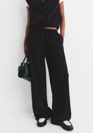 Person, die lockere schwarze Hose und eine schwarze ärmellose Weste über einem weißen Hemd trägt, eine schwarze Handtasche hält, mit schwarz-weißen Schuhen.