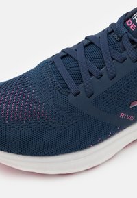 Zapatilla deportiva azul marino hecha de malla transpirable con detalles en rosa, que cuenta con una suela blanca, punta redondeada y cordones negros.