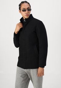 Svart ullblandingsfrakk med høy krage, frontglidelås og sidelommer. Modellen har på seg grå jeans og små solbriller.