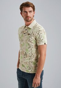 Polo a maniche corte in crema con un motivo floreale verde e marrone, caratterizzato da un colletto e tre bottoni neri.