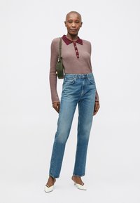 Mulher de pé, vestindo uma blusa de manga longa em malva com colarinho borgonha, jeans azuis, saltos brancos e uma mala de ombro verde-oliva.