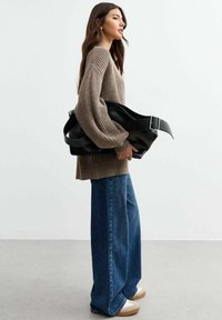 Una persona che indossa un maglione di lana marrone e jeans a gamba larga blu, tiene in mano una grande borsa in pelle nera con dettaglio a fibbia.