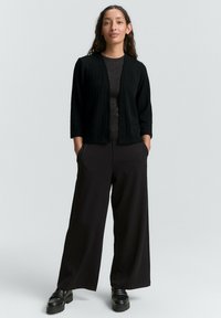 Černý žebrovaný cardigan přes tmavý top, spárovaný s širokými černými kalhotami a robustními černými botami. Modelka stojí s rukama v kapsách.