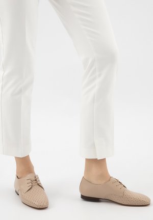 Person, die weiße Hose und beigen, perforierten Lederschnürschuhe trägt, steht vor einem weißen nahtlosen Hintergrund.