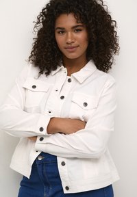 Chaqueta de mezclilla blanca con botones a presión, dos bolsillos en el pecho y mangas largas, combinada con jeans azules. Textura suave y diseño casual.