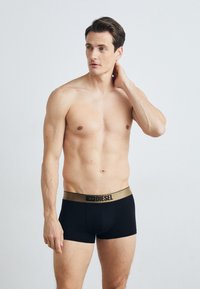 Black boxer briefs with a contrasting gold waistband featuring the brand logo. Model has a neutral pose and smooth skin texture.