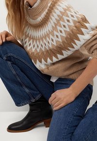 Muster gestrickter Pullover in Braun und Weiß mit hohem Kragen, kombiniert mit blauen Schlagjeans und schwarzen Ankle Boots mit einem kleinen Absatz.