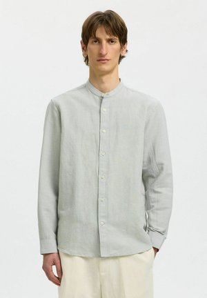 Homme aux cheveux bruns courts portant une chemise gris clair à col mandarin à manches longues et un pantalon crème, debout devant un fond clair uni.