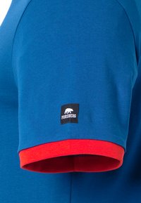 Blaues Kurzarmhemd mit rotem Bündchen. Verfügt über ein schwarzes Etikett mit einem Bärenlogo und dem Text "FORSGERG" am linken Arm. Glatte Textur.