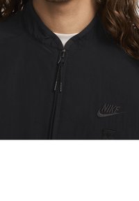 Svart lätt jacka med hög krage, dragkedja fram och broderad Nike-logotyp. Har en jämn textur och minimalistisk design.