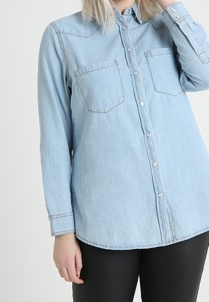 Kvinde iført en lyseblå denimskjorte med knapper, lange ærmer og to brystlommer, kombineret med sorte bukser.