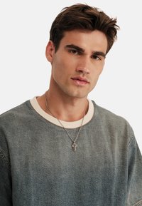 Sudadera de denim en azul desgastado con un cuello redondo crema y collar de cruz plateada. Textura suave; diseño casual con detalles de costura visibles.
