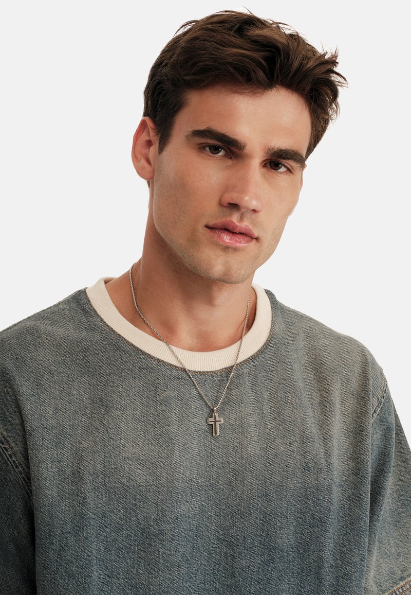 Sudadera de denim en azul desgastado con un cuello redondo crema y collar de cruz plateada. Textura suave; diseño casual con detalles de costura visibles.