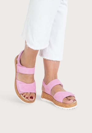 Füße in rosa Wildleder-Keilabsatzsandalen mit Korksohlen und weißen eingefassten Sohlen, kombiniert mit weißen, gekürzten Hosen mit ausgefransten Säumen.