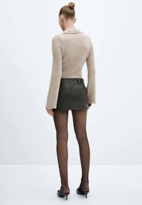 Femme avec les cheveux en chignon portant un pull beige à côtes, une mini-jupe en cuir noir, des collants noirs transparents et des chaussures à talons kitten noires, de dos.