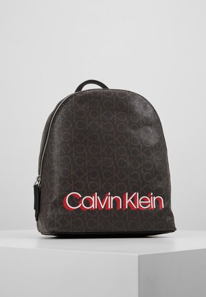 Mørkebrun Calvin Klein rygsæk med gentagende "ck" monogrammønster og stort hvidt og rødt Calvin Klein-logo foran, stående på en hvid overflade.