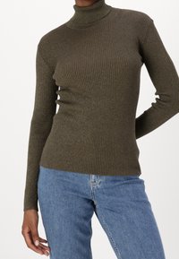 Personne portant un pull col roulé côtelé vert olive et un jean bleu, avec une main reposant sur la hanche et un fond neutre.