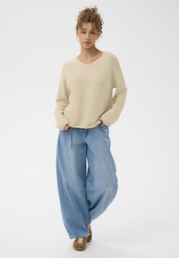 Pull beige à manches longues et col en V associé à un jean en denim bleu clair et ample, ainsi qu'à des chaussures marron. Look décontracté avec une silhouette relaxée.