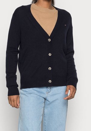 Persoon die een zwart button-up vest draagt over een beige trui en lichtblauwe spijkerbroek tegen een effen witte achtergrond.