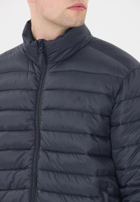 Mörkblå puffjacka med hög krage, horisontellt quiltad design, zipfrontslutning och en diskret logotyp på bröstet. Slät textur.