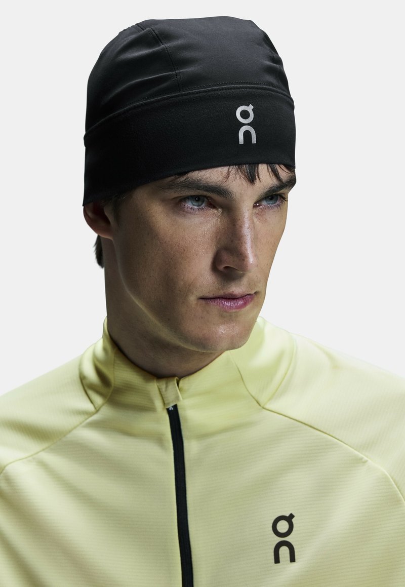 Gorro de running negro con una textura suave y elástica. Presenta un logo y un diseño minimalista. Se lleva con una chaqueta amarilla clara.