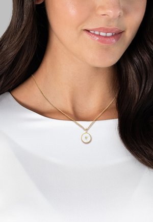 Collana con pendente in oro caratterizzata da un charm circolare con un motivo a raggiera testurizzato, indossata su una delicata catena d'oro, su sfondo di una maglietta bianca.
