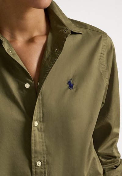 Közelkép egy személyről, aki olívazöld gombos inget visel, melynek mellkasán egy kis sötétkék hímzett logó látható.