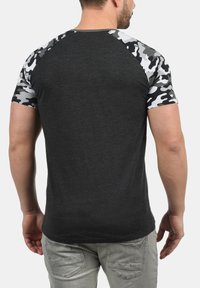 Black t-shirt with camo-patterned raglan sleeves, fitted design, short sleeves, and a smooth texture. Worn with light-colored pants.