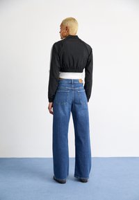 Veste noire courte avec des rayures blanches, associée à un jean bleu en denim à jambes larges. La tenue présente une taille ajustée et deux poches arrières.