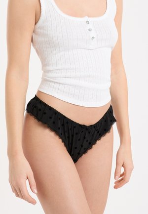 Schwarze Pantys mit weißen Polka Dots, Spitzenbesatz, Schleifendetail und gerafftem Bund. Hergestellt aus weichem Stoff, mit niedrig sitzendem Schnitt und verspieltem Muster.