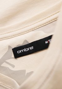 Camiseta de tela beige con una textura suave, que presenta una etiqueta negra que dice "ombre" y un sutil patrón de camuflaje claro.