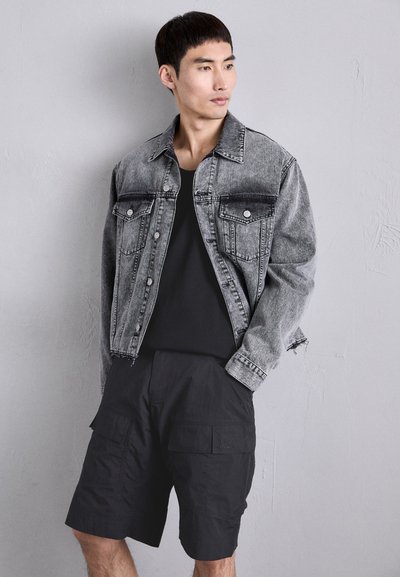 Veste en denim noire et grise avec deux poches poitrine et fermeture à boutons ; associée à un short cargo noir avec des poches avant.