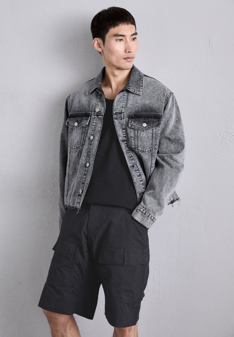 Veste en denim noire et grise avec deux poches poitrine et fermeture à boutons ; associée à un short cargo noir avec des poches avant.