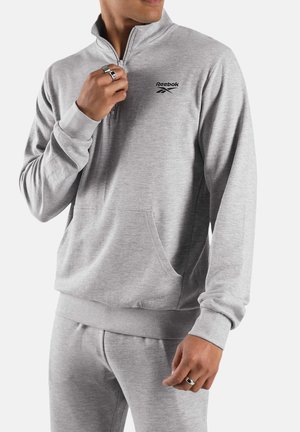 Hombre con sudadera Reebok gris claro con cremallera hasta un cuarto y pantalones deportivos a juego, ajustando la cremallera con la mano izquierda, anillos visibles en los dedos.