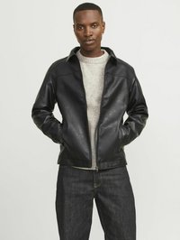 Chaqueta de cuero negra con cierre de cremallera y cuello, que presenta una textura suave, situada sobre un fondo neutro. Lleva puesta una sudadera gris.