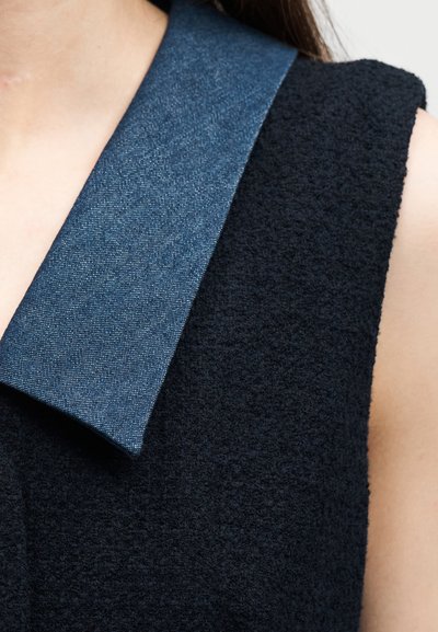 Κοντινό πλάνο ενός ατόμου που φοράει ένα μαύρο υφασμάτινο ρούχο χωρίς μανίκια με ένα φαρδύ, επίπεδο μπλε γιακά πάνω στον ώμο.