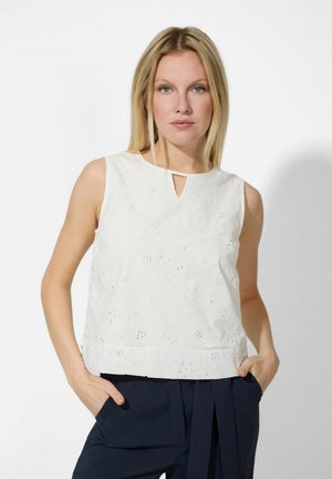 Top sans manches blanc avec broderies florales, décolleté en forme de goutte et ourlet droit. Porté avec un pantalon foncé. Texture lisse.