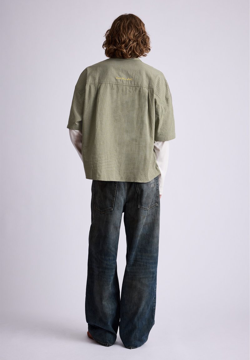 Personne aux cheveux bouclés portant une chemise à manches courtes verte à carreaux sur une chemise blanche à manches longues et un jean large foncé, debout, dos à l’objectif.