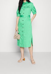 Grünes Hemdkleid mit kurzen Ärmeln, tailliertem Design und seitlichen Schlitzen. Es hat schwarze Knöpfe und eine glatte Stoffoberfläche.
