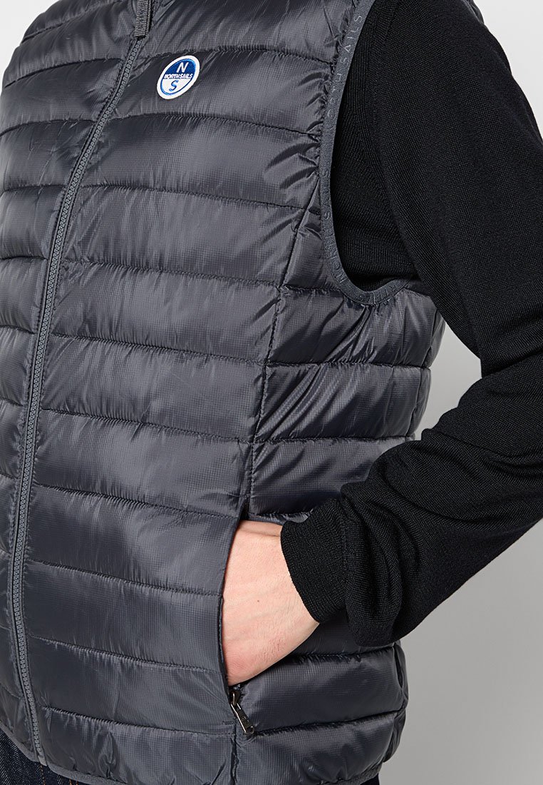 Gilet grigio imbottito con motivo trapuntato orizzontale, cerniera frontale e toppa con logo. Indossato sopra una maglietta nera a maniche lunghe, mani nelle tasche.