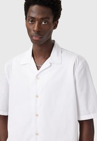 Camisa de botones de manga corta blanca confeccionada con un tejido ligero, presenta un cuello clásico y sutiles detalles en los botones. Textura suave.