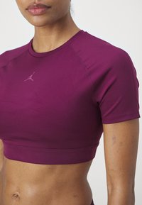 Žena v těsném fialovém crop topu s malým logem Jumpman na hrudi, odhalující břicho a paži na jednobarevném pozadí.