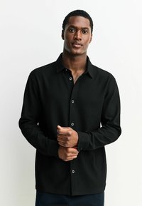 Melnā ribota krekla ar apvalku, kuram ir melnas pogas un garas piedurknes. Audums izskatās teksturēts un nedaudz elastīgs.