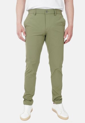 Uomo che indossa pantaloni slim-fit verde oliva, sneakers bianche basse e una camicia bianca, in piedi contro uno sfondo semplice.