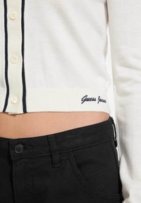 Witte gebreide cardigan met zwarte afwerking en knoopsluiting, voorzien van "Guess Jeans" geborduurd in marineblauw aan de zoom, gecombineerd met zwarte jeans.