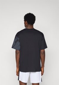 Sort bomulds T-shirt med korte ærmer, der har et tekstureret mønster i mørkegrå på venstre ærme og en afslappet pasform, båret med hvide shorts.