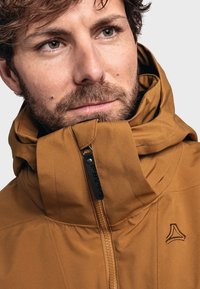 Brown zip-up jacket with a high collar and logo. Features a sleek design, smooth texture, and visible stitching details.
