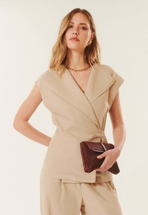 Femme aux longs cheveux portant un haut cache-cœur beige sans manches et un pantalon, tenant une pochette marron foncé texturée, accessoirisée avec des bijoux en or.